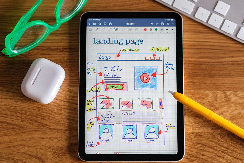 A tablet with a hand-drawn sketch of a webpage on a wooden table. On the top left is a pair of neon green glasses and part of a keyboard in the top right.